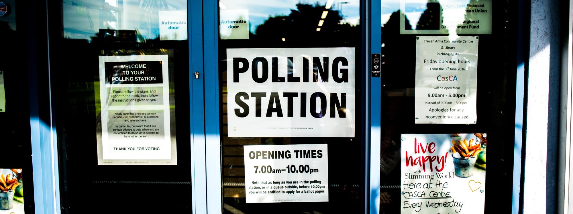 Eine Glastür mit vershiedenen Informationsschildern, in der Mitte ein großes Schild mit der Aufschrift "Polling Station"