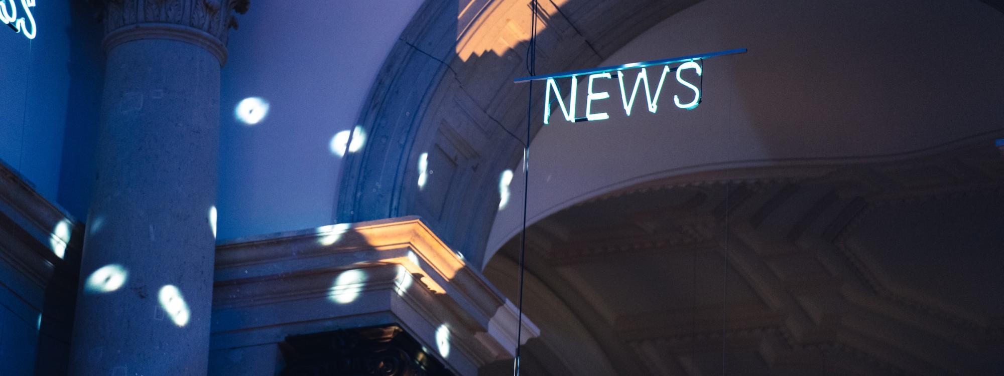 Ein Deckenbogen in einem alten Gebäude, vielleicht ein Museum, nur partiell beleuchtet, unter der Decke hängt ein Neonlicht, das das Wort "NEWS" formt.